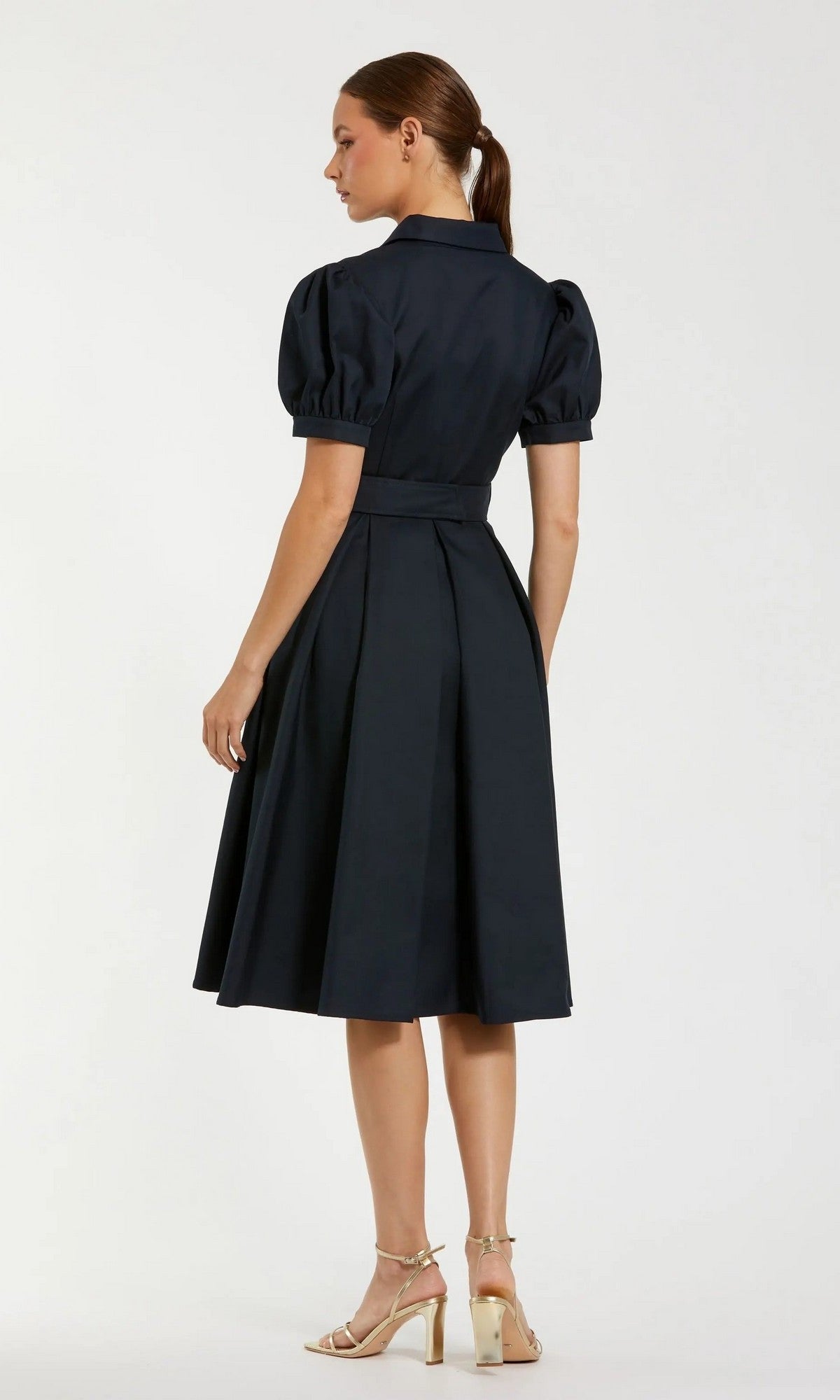 A woman stands with her back to the camera against a plain background, wearing the Mac Duggal 69097 Knee Length Belted Shirt Dress in navy blue, which features a collared neckline, short sleeves, and an A-line skirt. She pairs the dress with gold heeled sandals.